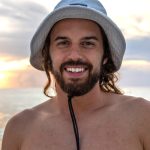 Young man with a beard and a sunhat standing in front of the ocean during sunset.